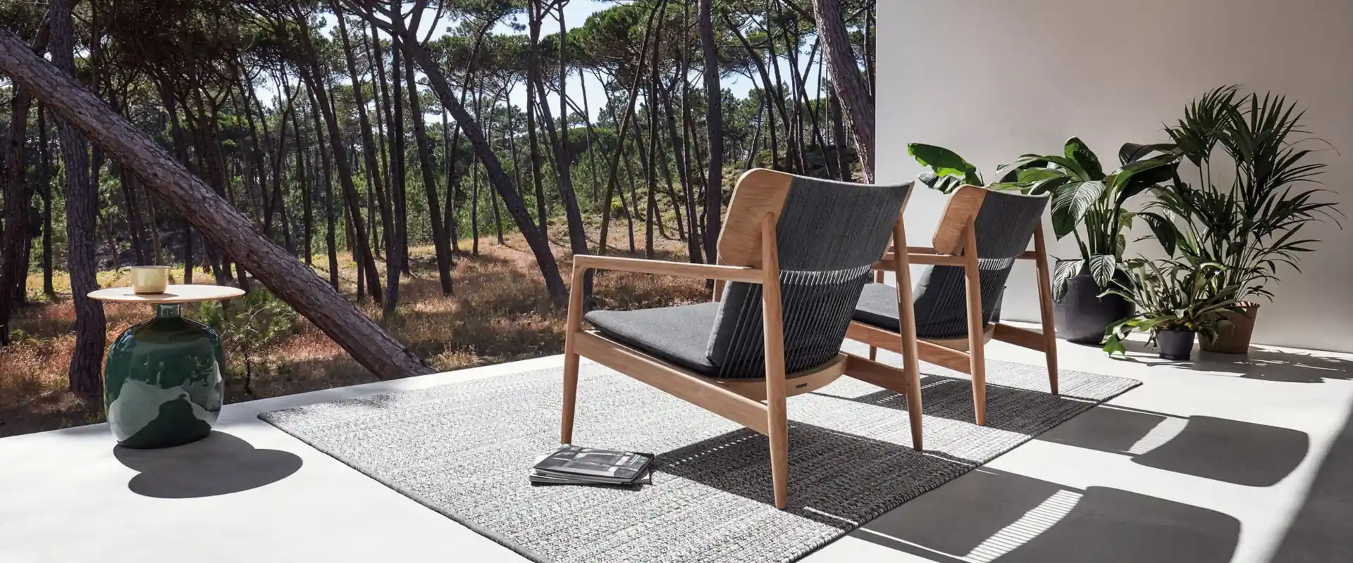 Gloster Archi teak lounge chairs with dark woven seats on a rug, on a terrace opening onto a pine wood