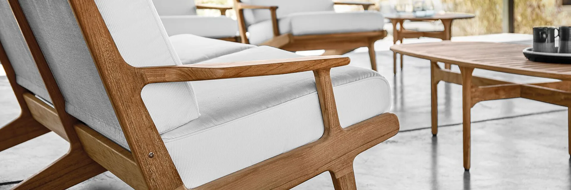 Close view of the teak frame and white cushions of a Gloster Bay lounge chair beside a teak coffee table, in bright sunlight
