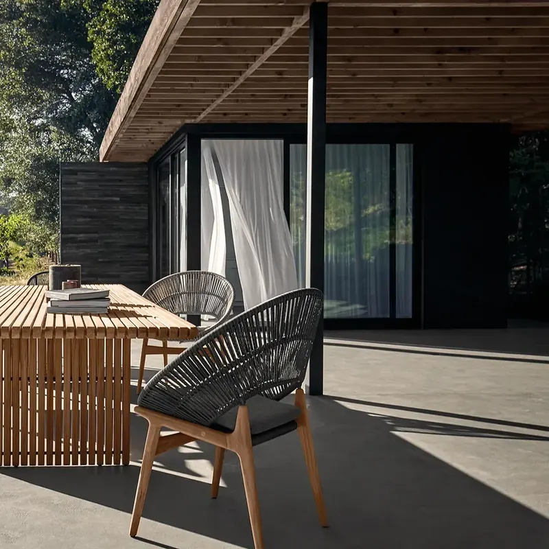 Gloster Bora dining chair in dark rope at a slatted teak dining table on a terrace beside a dark timber house