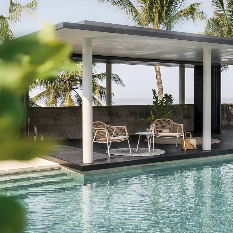 Gloster Fresco lounge chairs and a small table under a white pavilion set in a turquoise pool, a palm tree behind