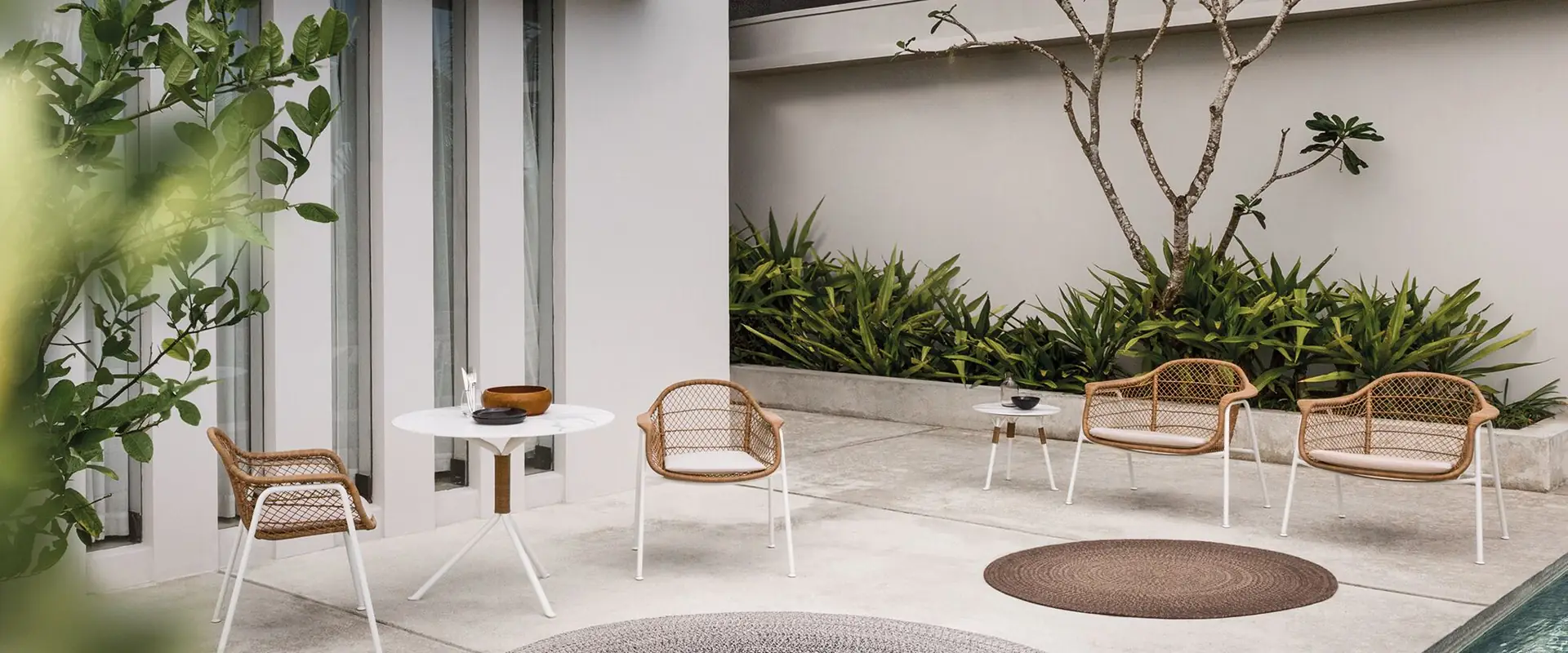 Gloster Fresco woven chairs around small tables on the patio of a white house, with a round rug and tropical planting