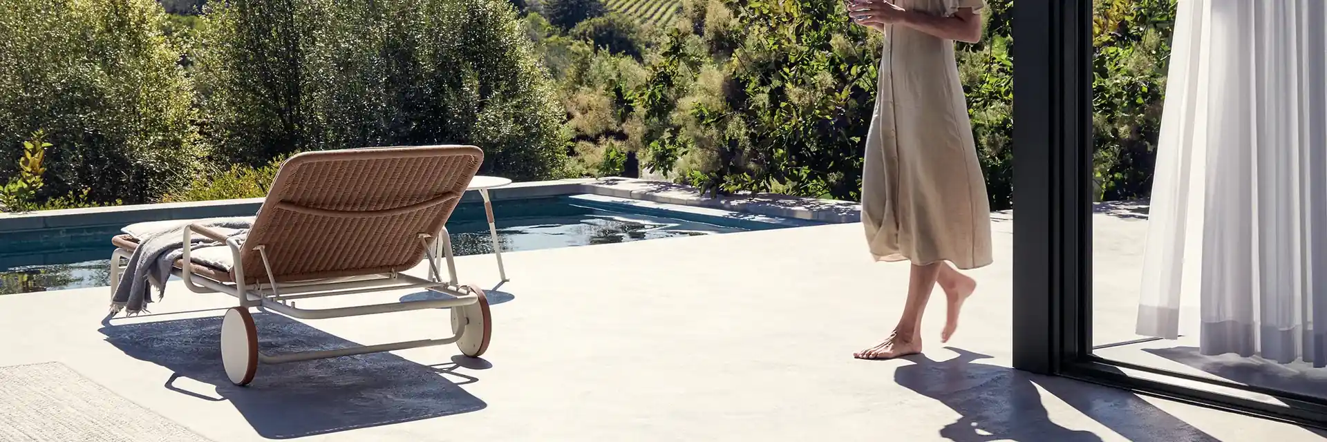 Gloster Fresco woven sun lounger on a pool terrace, with a woman walking past and wooded hills behind