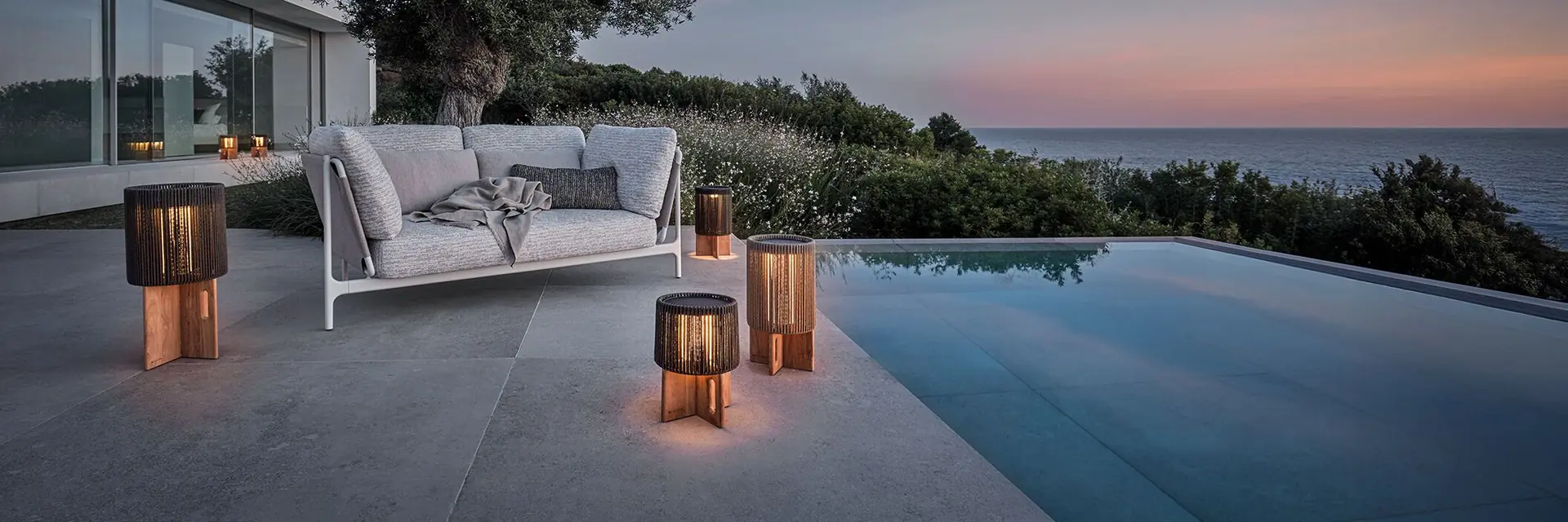 Gloster Grand Sail sofa with lanterns on a terrace beside an infinity pool at dusk, with the sea beyond