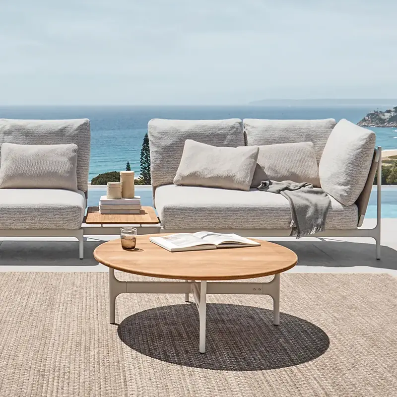 Gloster Grand Sail modular sofa with a round teak coffee table on a woven rug, the sea on the horizon
