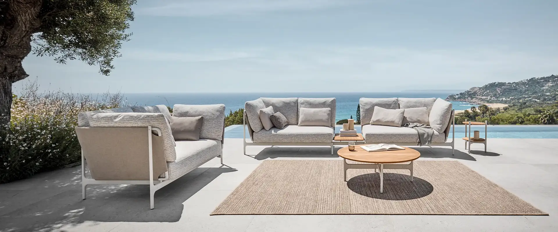 Gloster Grand Sail sofas with pale cushions around a round table on a terrace overlooking the sea