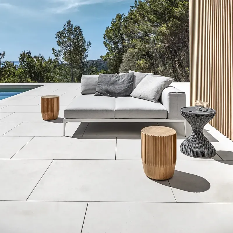 Gloster Grid sofa with gray cushions and round teak side tables on a white terrace beside a slatted wooden wall