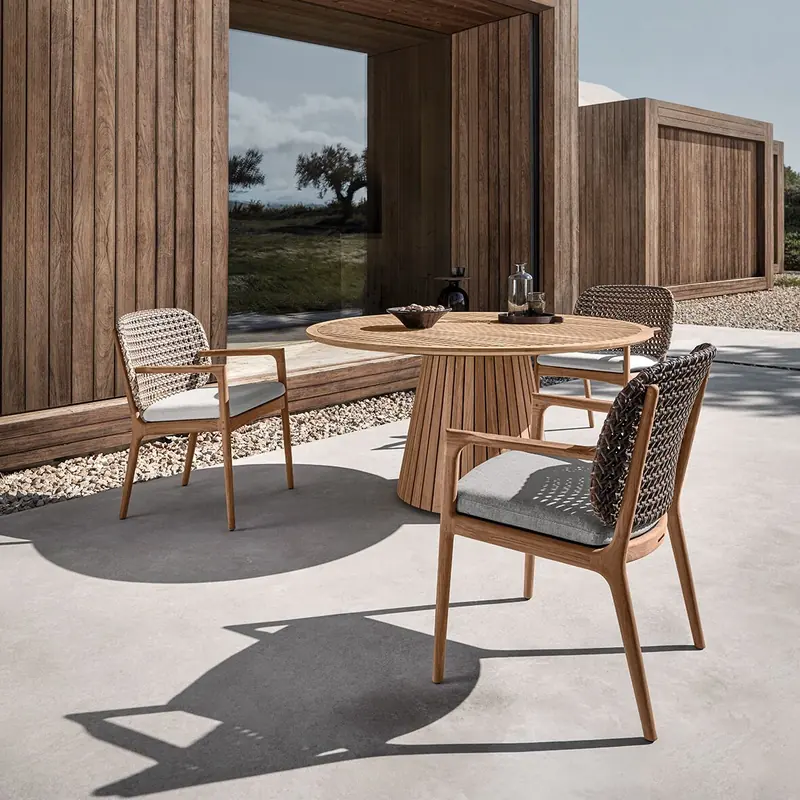 Gloster Kay dining chairs with arms, in woven wicker and teak, around a round teak table on a concrete terrace beside a timber-clad house