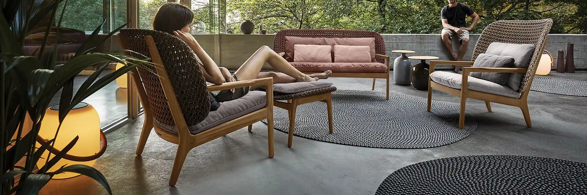 Gloster Kay high-back lounge chairs with woven shells and teak frames on a covered concrete terrace open to a forest, with a woman reading in one of them