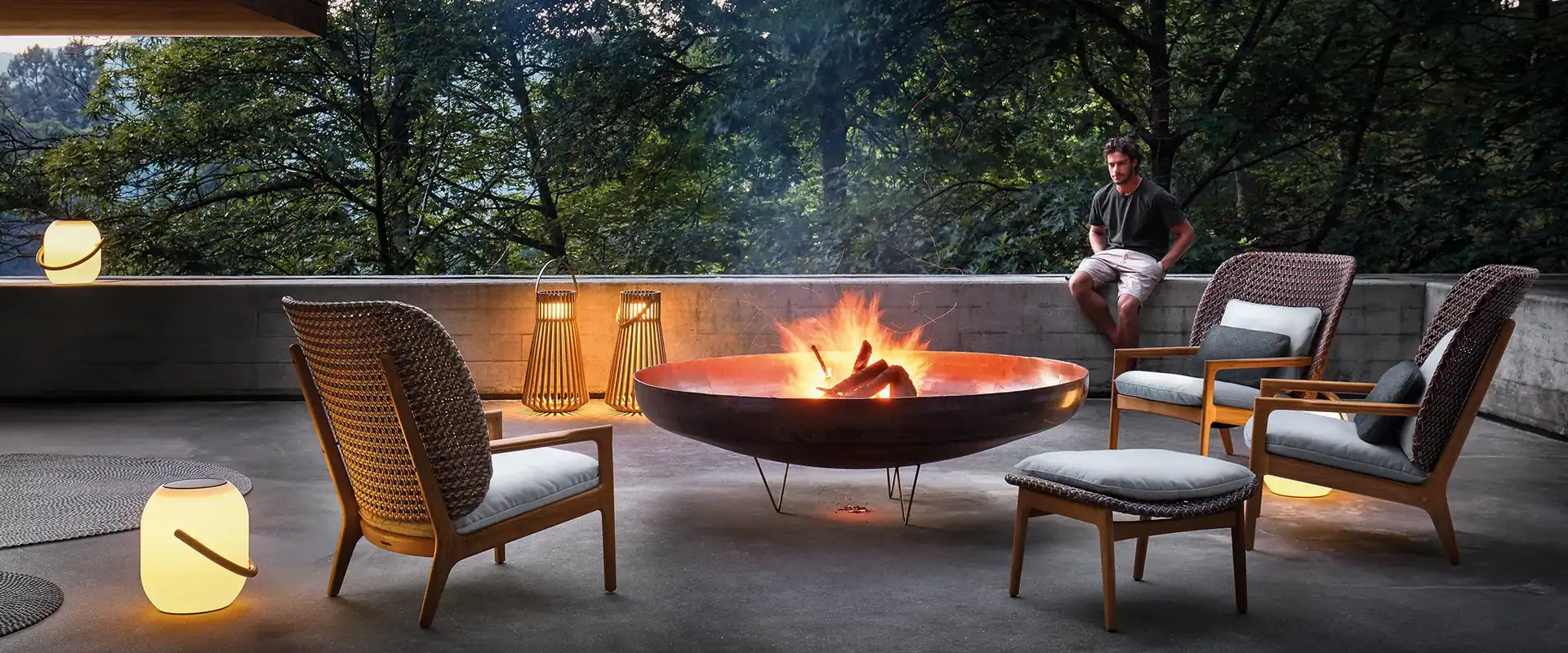 Gloster Kay woven lounge chairs with teak frames and a footstool around a fire bowl on a concrete terrace at dusk, with lanterns and trees behind