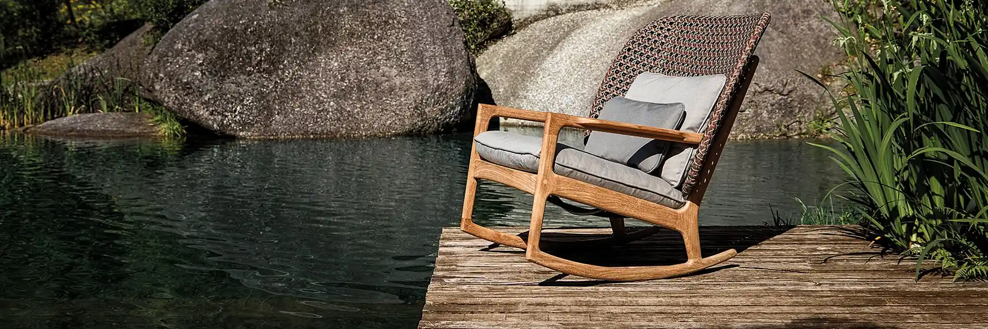 Gloster Kay rocking chair with a woven high back and teak frame on a timber deck beside a natural pool with granite boulders