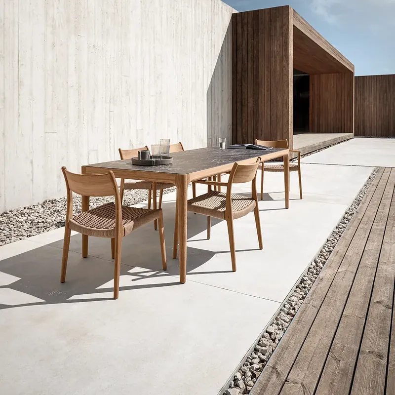 Gloster Lima teak dining chairs around a table on a pale terrace along a tall concrete wall
