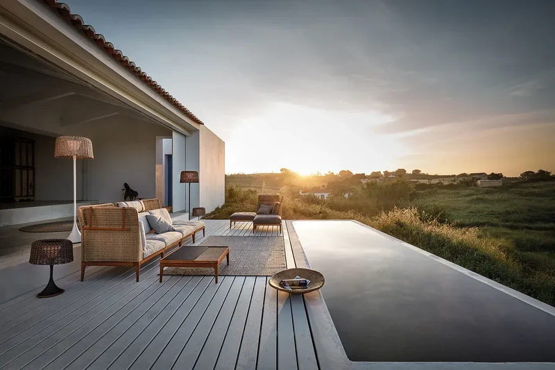 Gloster Lima teak sofa and coffee table on a timber deck beside a reflecting pool at sunset, with a floor lamp and a white house