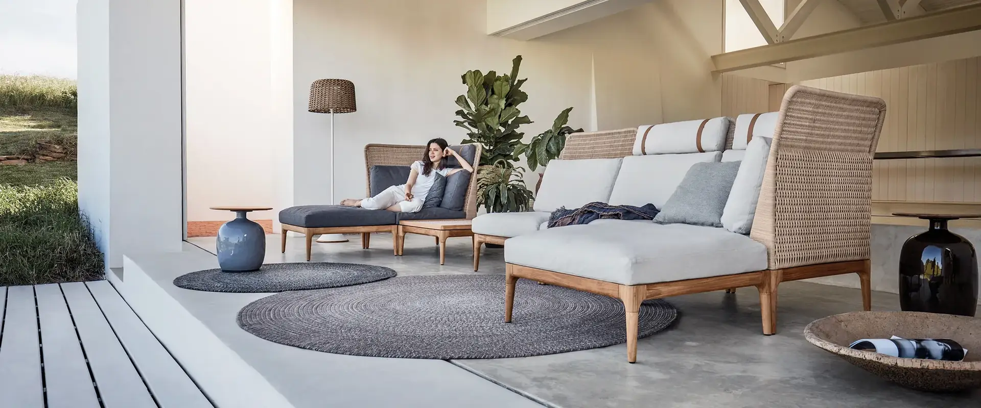 Gloster Lima high-back sofa in woven wicker with pale cushions on a porch with white beams, a woman resting on a daybed behind