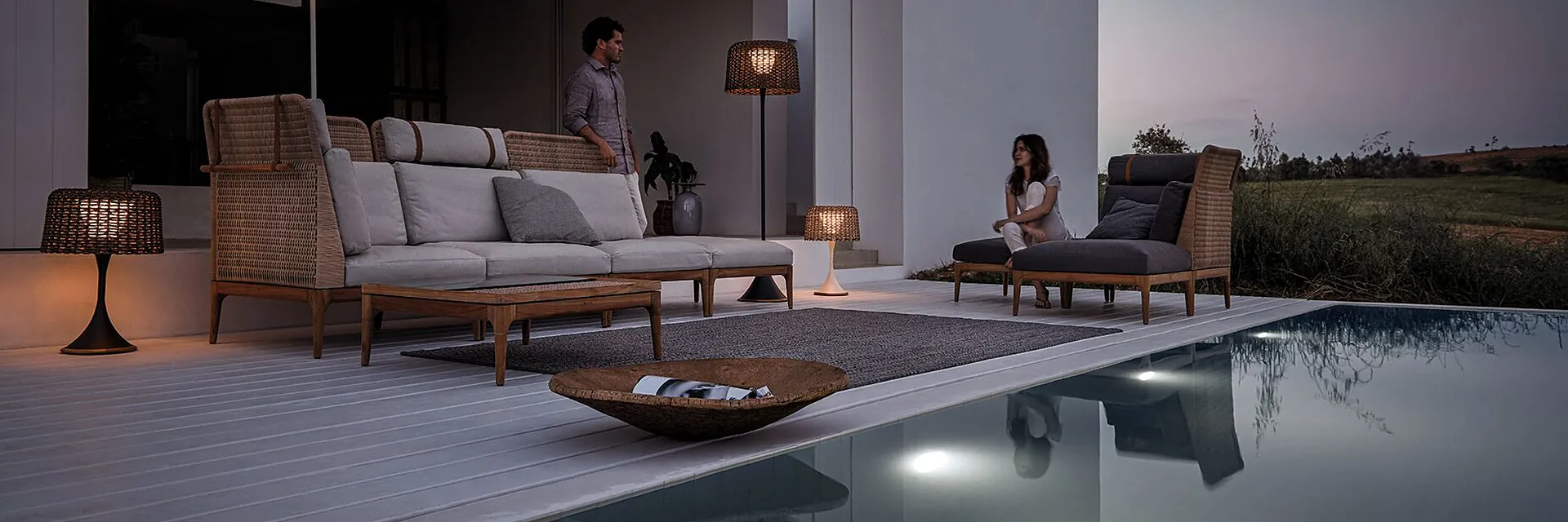 Gloster Lima sofa and lounger with woven teak frames beside a dark pool at dusk, with floor lamps and two people on the terrace of a white house