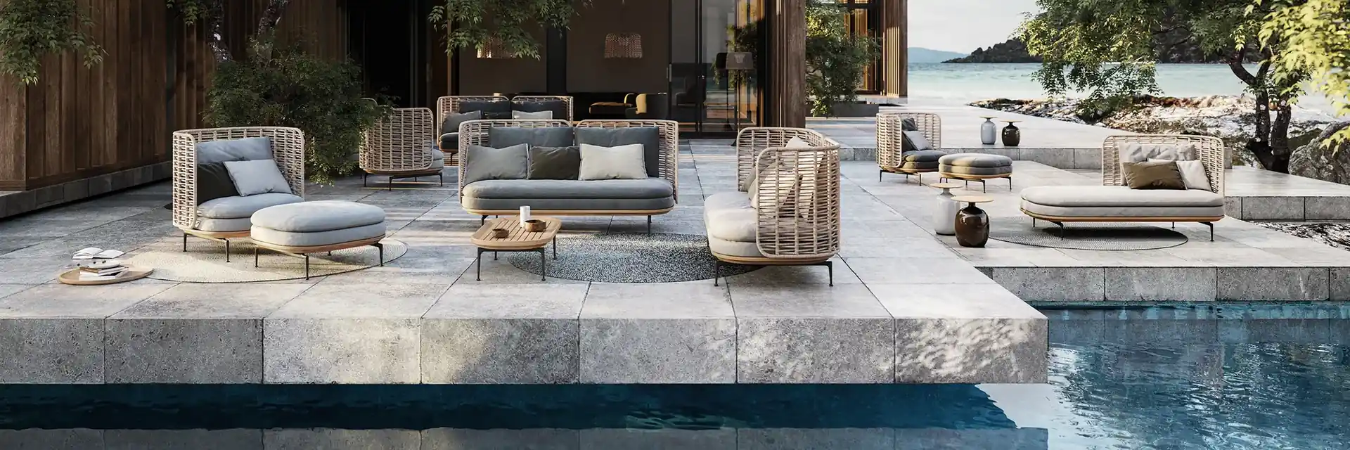 Gloster woven lounge chairs and sofas on a stone terrace beside a pool, with pine trees and a sea cove behind