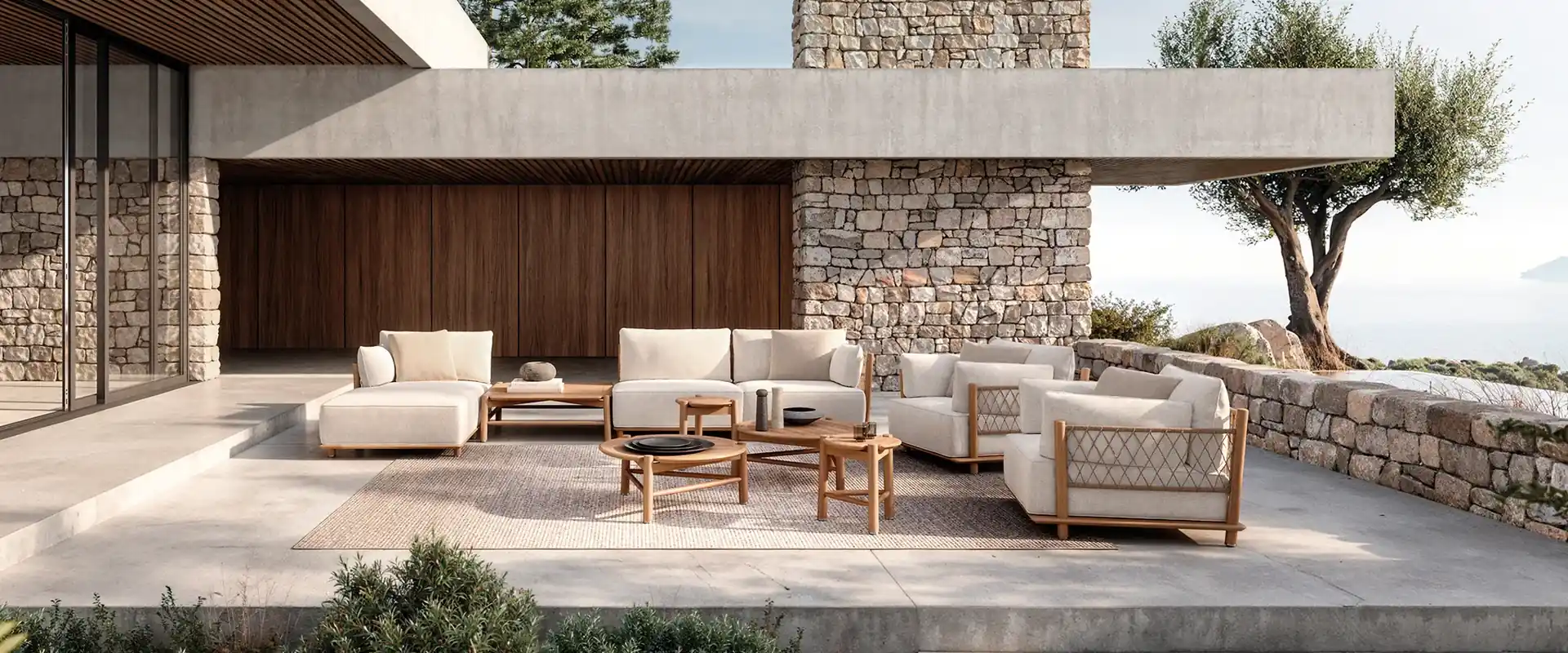 Gloster teak sofas and armchairs with cream cushions on the terrace of a villa with stone walls, an olive tree and the sea beyond