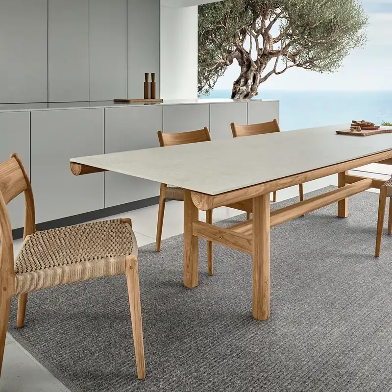 Gloster Rail teak dining table and woven chairs on a rug on a terrace, with an olive tree and the sea behind