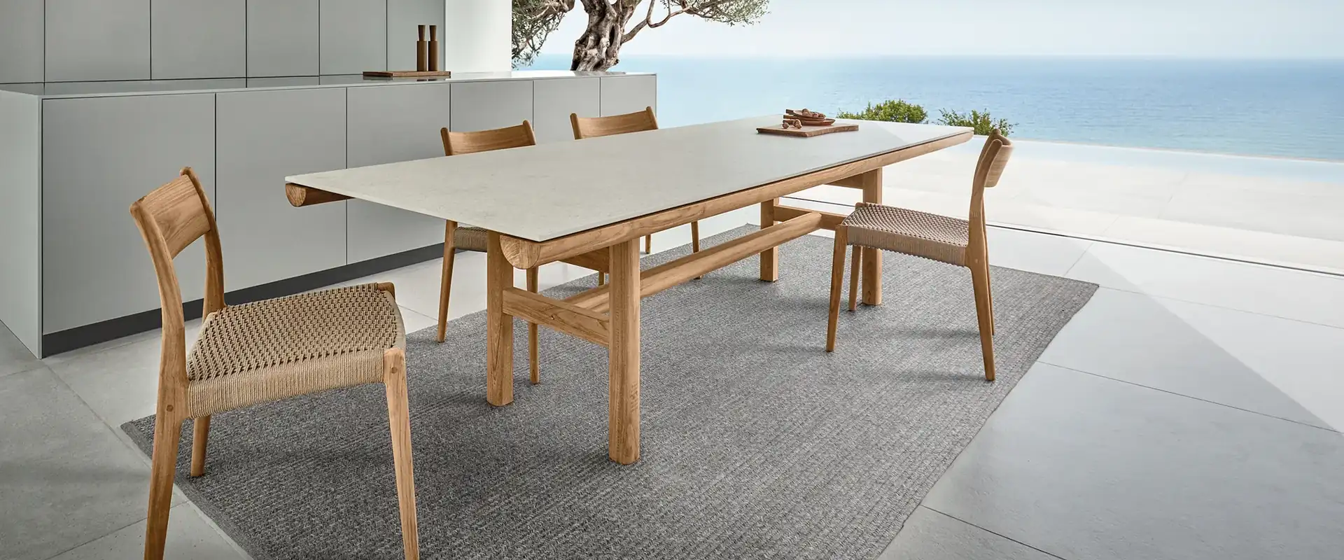 Gloster Rail teak dining table and woven chairs on a rug on a terrace, with an olive tree and the sea behind