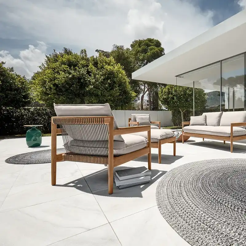 Gloster Saranac teak lounge chair and sofa with pale cushions on a white terrace in front of a glass-walled house