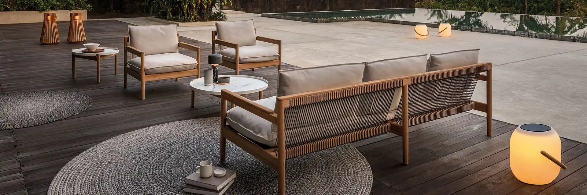 Gloster Saranac sofa and lounge chairs on a timber deck at dusk, with a lantern and palm trees behind