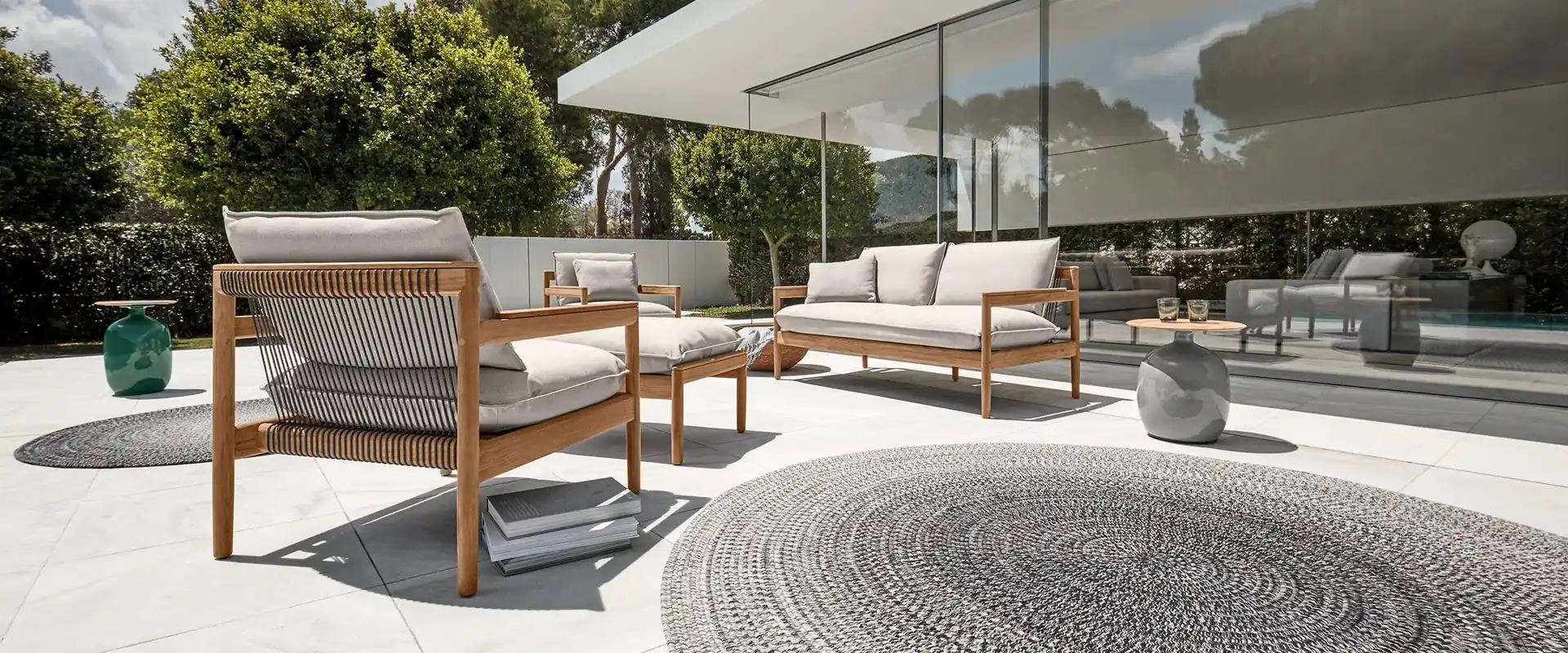 Gloster Saranac teak lounge chairs and sofa with white cushions on a round rug beside a glass pavilion