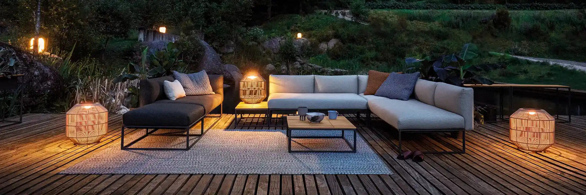 Gloster modular sofa and coffee table on a wooden deck at night, lit by lanterns, with a lit pavilion in the trees behind