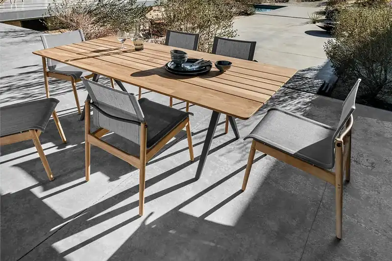 Gloster Split teak dining table with Sway teak armchairs on a covered terrace, with a pool house and mountains behind