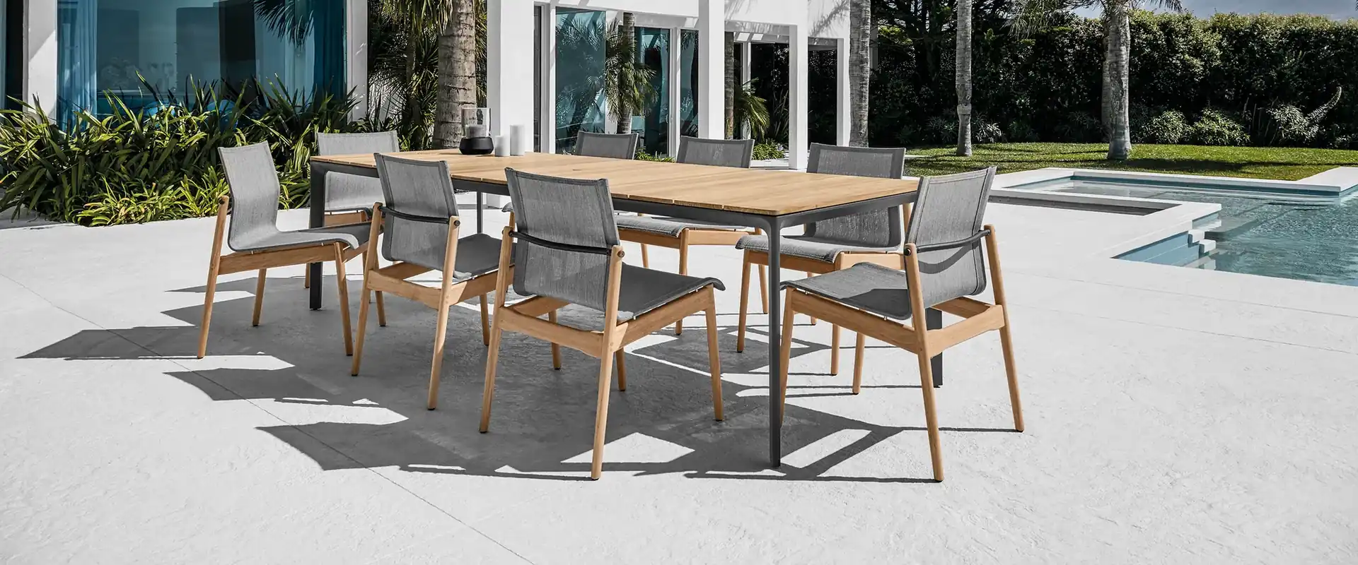 Gloster Sway teak dining chairs with sling seats around a long table on a white terrace, a white villa and palm trees behind