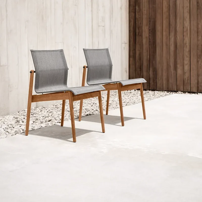Two Gloster Sway teak chairs with pale sling seats on a white floor, against a wall of concrete and dark timber