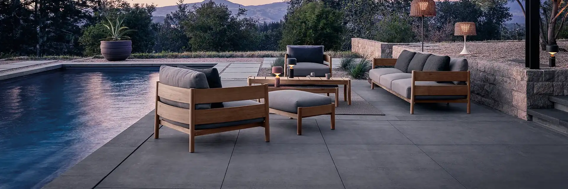 Gloster teak sofas and armchairs with dark cushions on a stone terrace beside a pool at dusk, hills in the distance