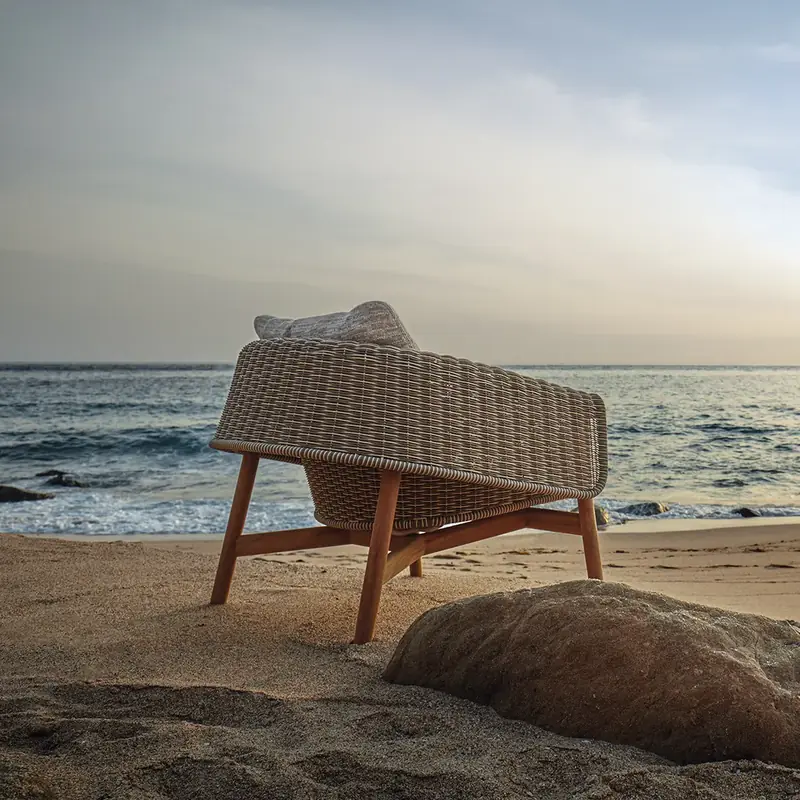 Gloster Wrap lounge chair with a woven wicker back standing on the sand among rocks, facing the sea at sunset