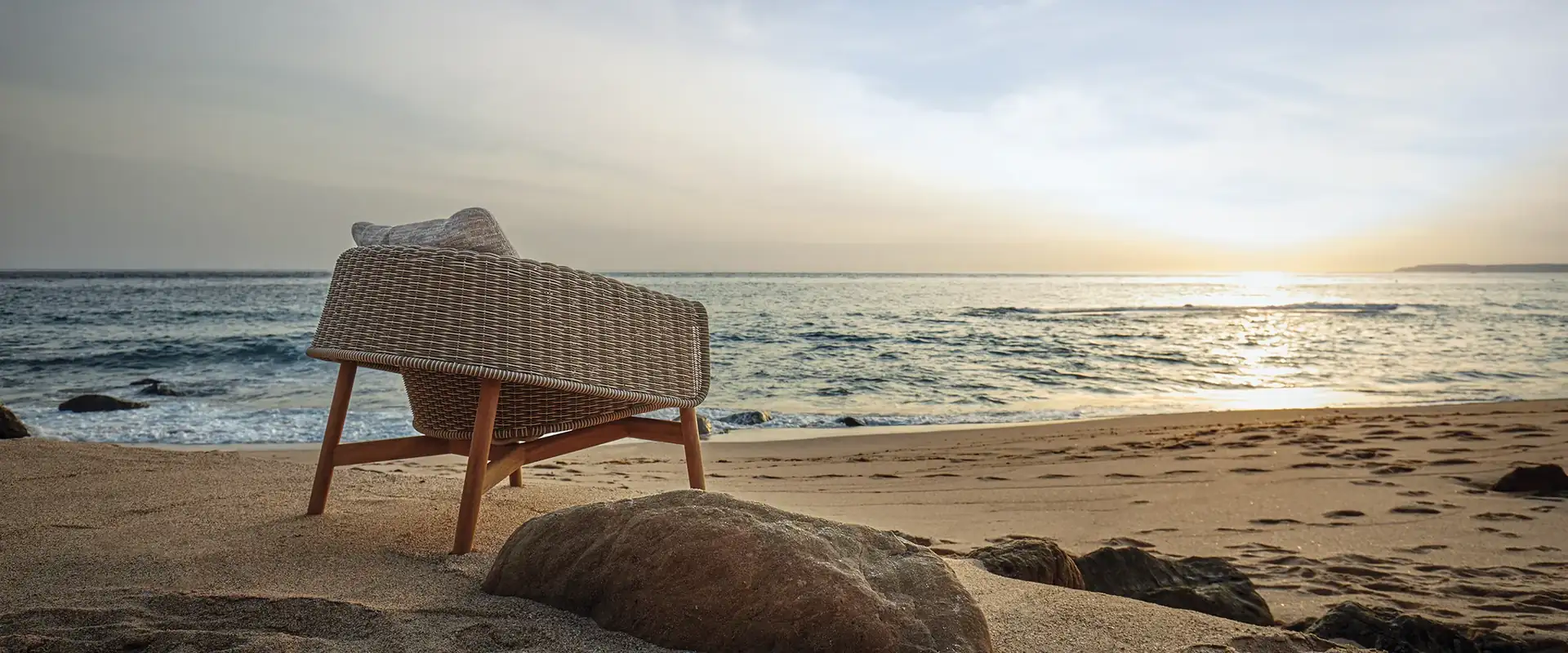 Gloster Wrap lounge chair with a woven wicker back standing on the sand among rocks, facing the sea at sunset
