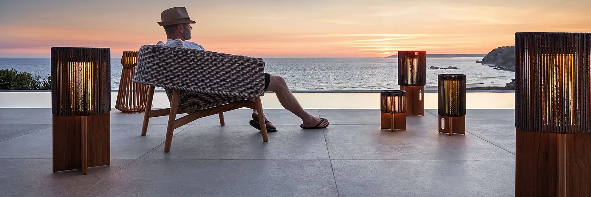 Man sitting in a Gloster Wrap lounge chair on a terrace by the sea at sunset, surrounded by timber lanterns