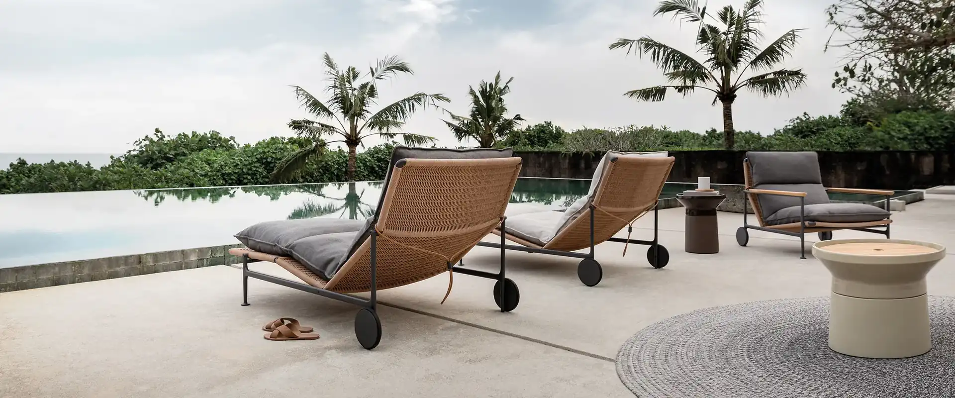 Gloster Zenith sun loungers with tan slings on a terrace beside an infinity pool, with palm trees and a cloudy sky