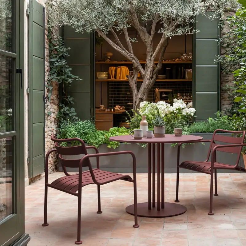 Kettal Atlantic aluminum armchairs in burgundy around a round bistro table on a terracotta-tiled patio with an olive tree and potted plants