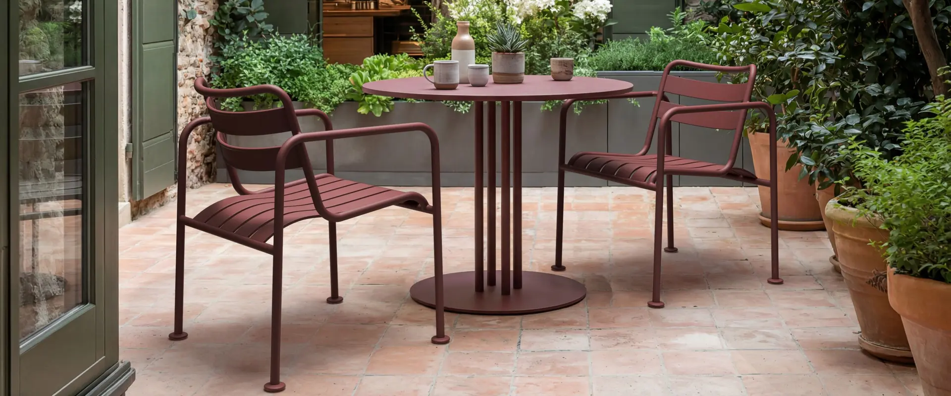 Kettal Atlantic aluminum armchairs in burgundy around a round bistro table on a terracotta-tiled patio with an olive tree and potted plants