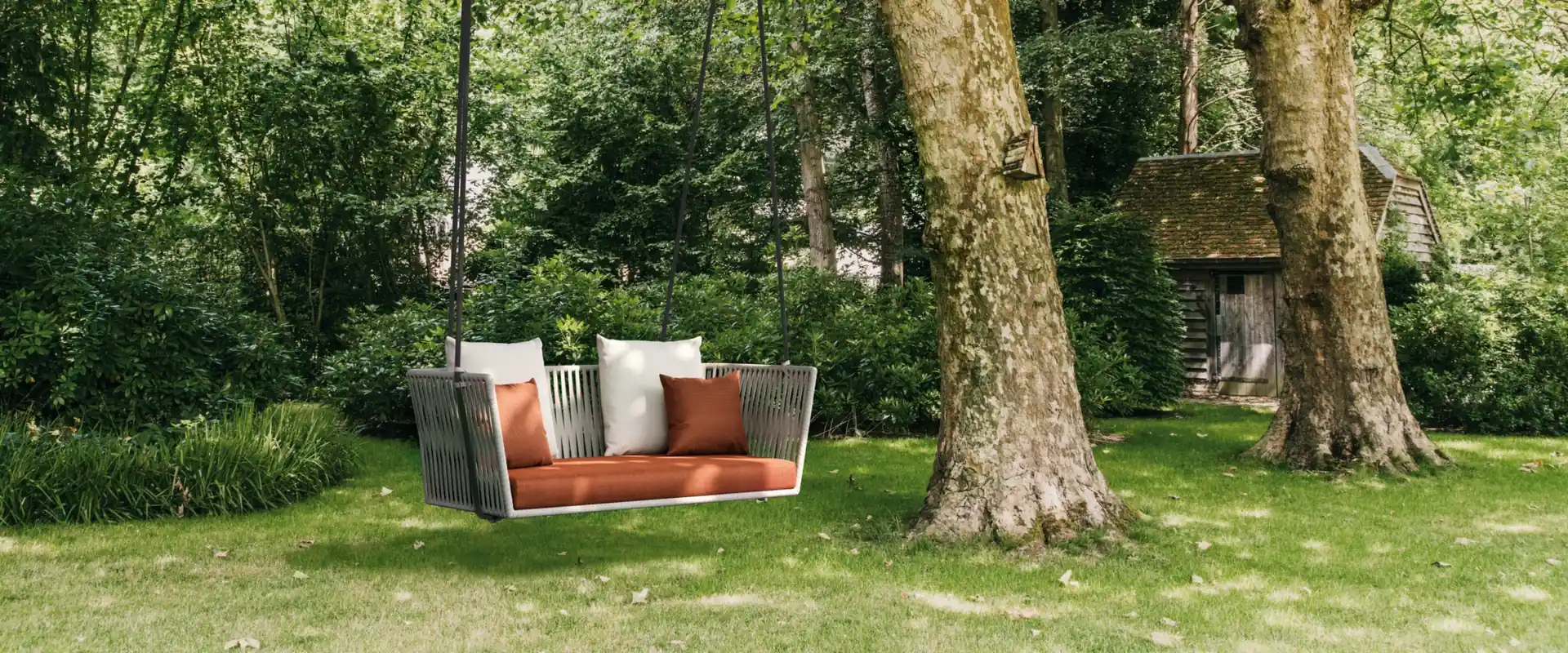 Kettal Bitta two-seater hanging sofa with orange cushions suspended from a large tree on a green lawn