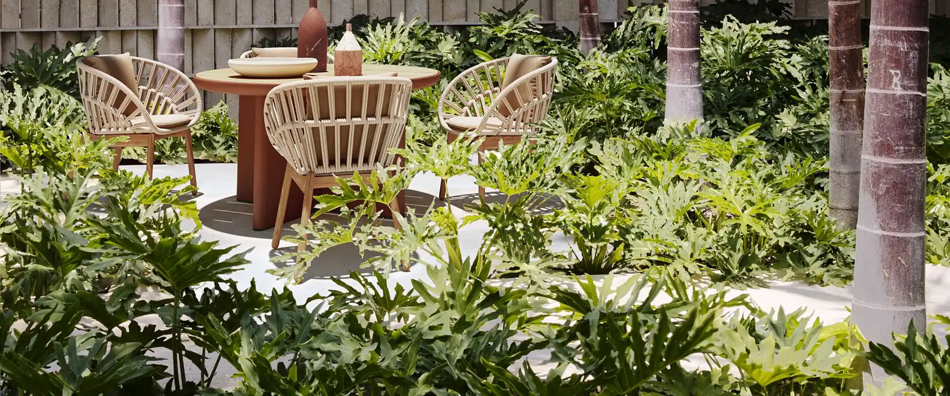 Kettal Cala rope armchairs around a round terracotta table in a garden of palm trees and tropical plants, in front of a lattice wall