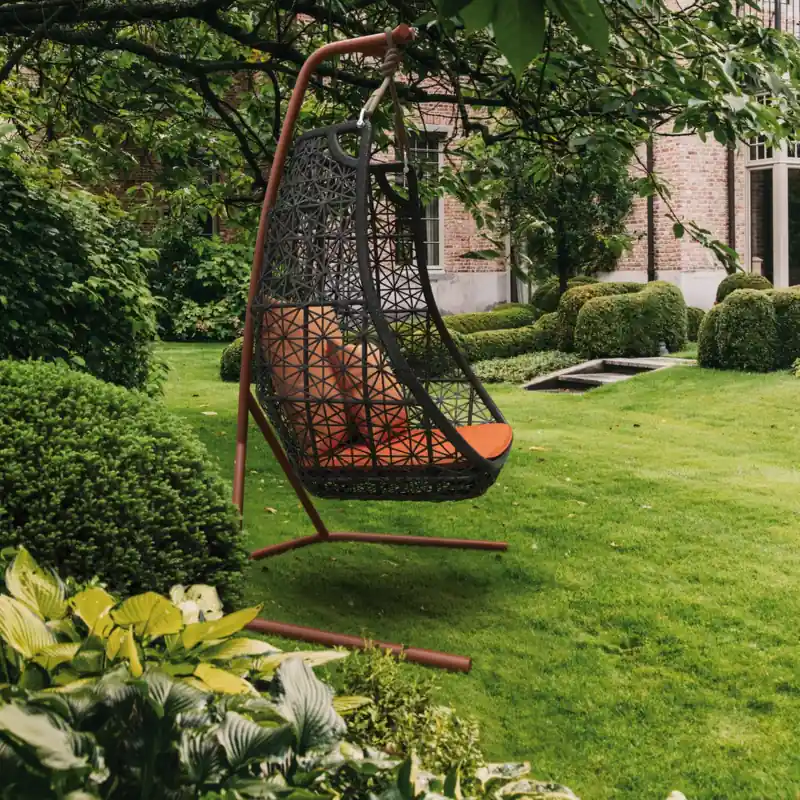 Kettal Maia hanging egg chair with orange cushions on its stand, on a lawn under a tree with a brick house behind