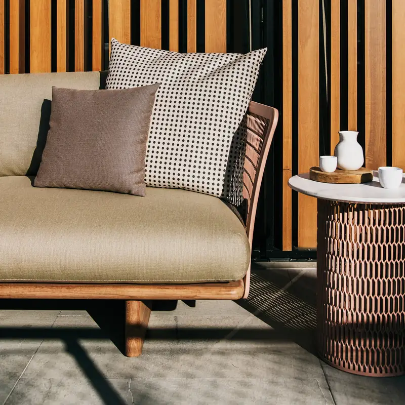 Corner of a Kettal Mesh sofa with beige cushions and a round side table, in the striped shade of a slatted wooden screen