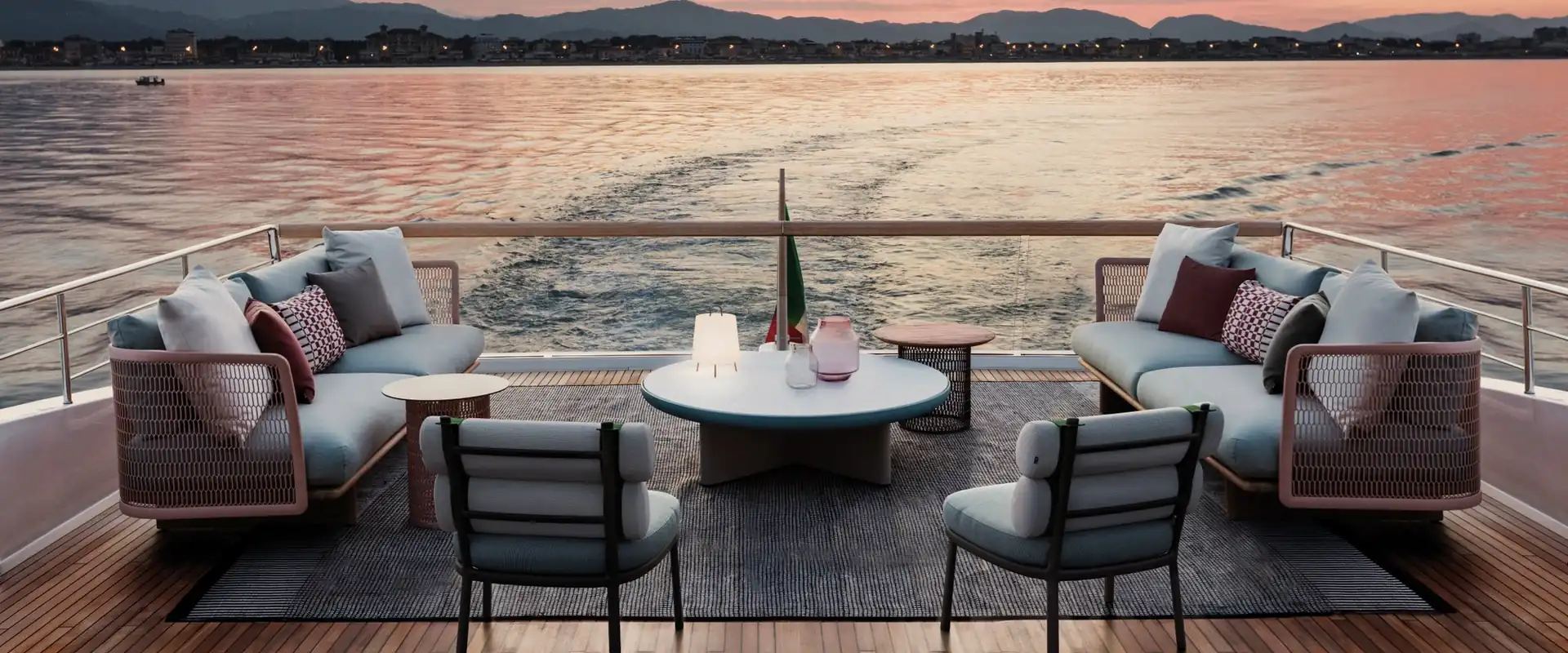 Kettal Mesh sofas and armchairs around round coffee tables on the teak deck of a yacht, with the sea and a pink sunset sky