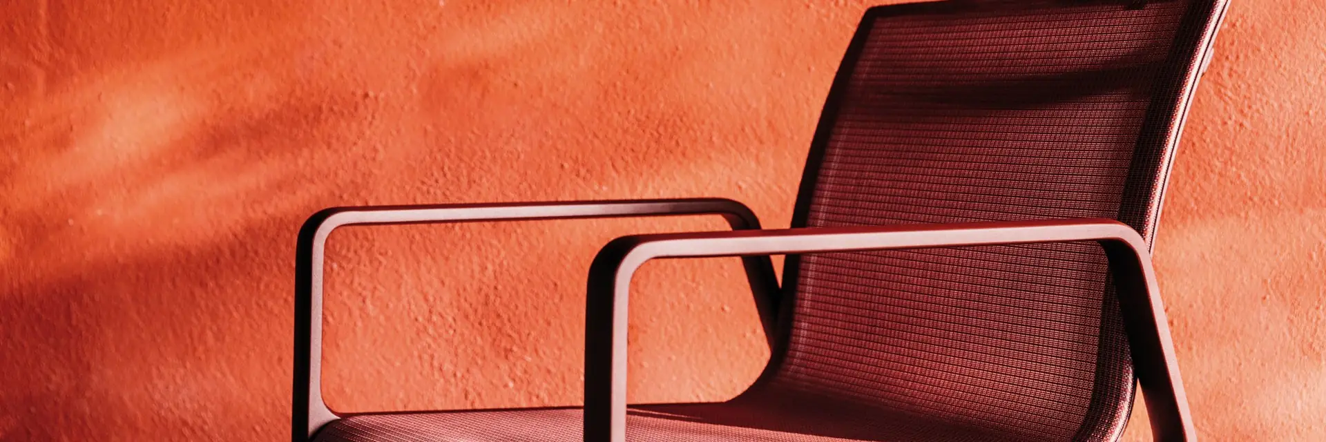Kettal Park Life aluminium armchair with a burgundy mesh seat against a terracotta wall in low sunlight