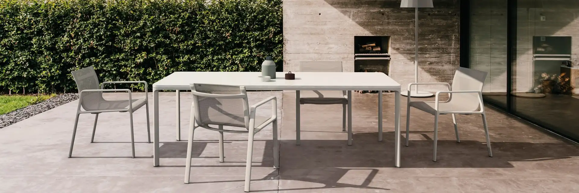Kettal Park Life aluminium dining table and chairs on the covered terrace of a concrete house, with a hedge and meadow behind