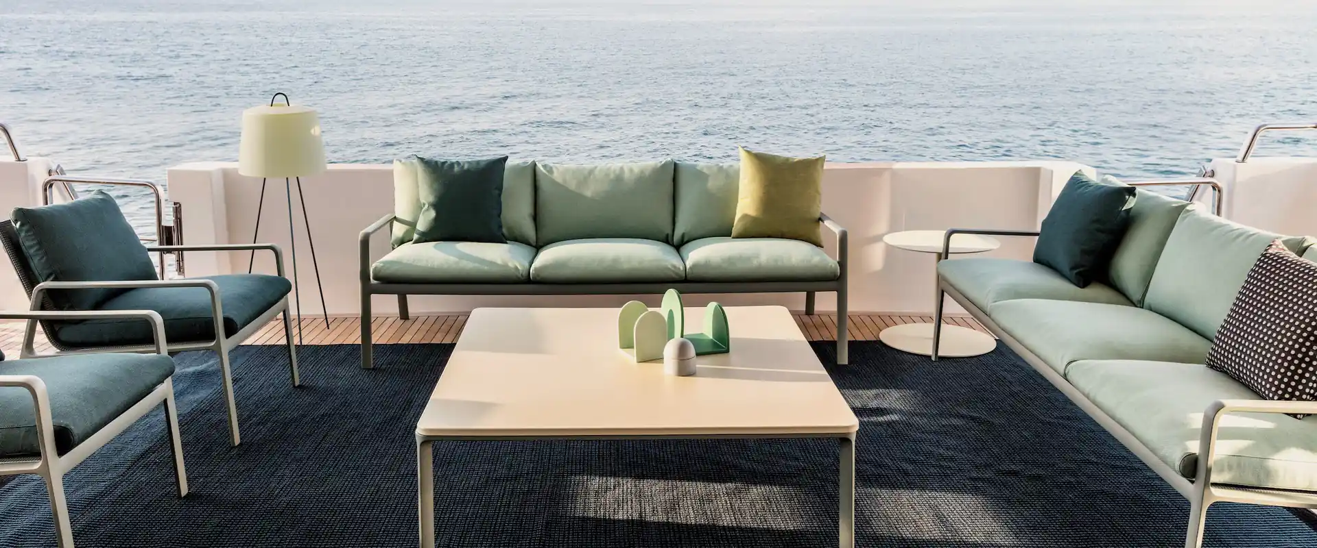 Kettal Park Life sofas and armchairs with green and blue cushions around a low table on the deck of a yacht, with the open sea behind
