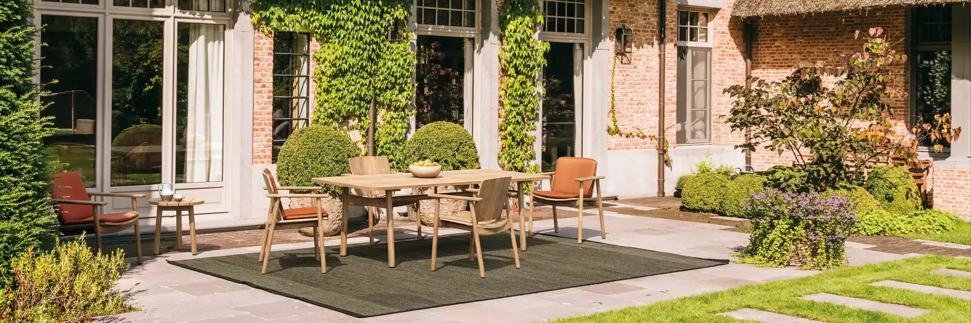 Kettal Riva teak dining table and chairs on a rug in the garden of an ivy-clad brick house