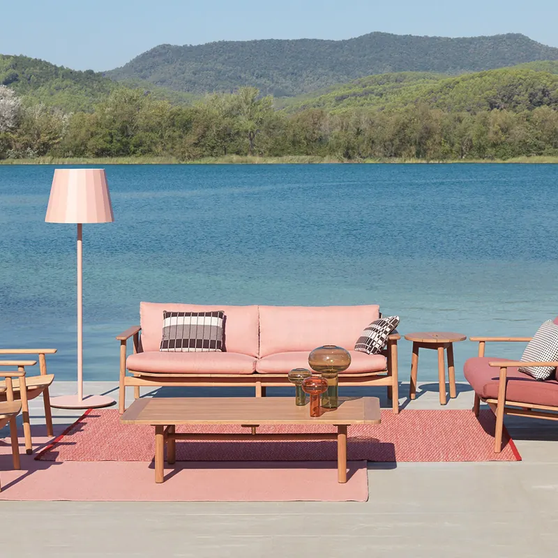 Kettal Riva teak sofa and armchairs with pink cushions on a red rug at the edge of a lake, with green hills behind