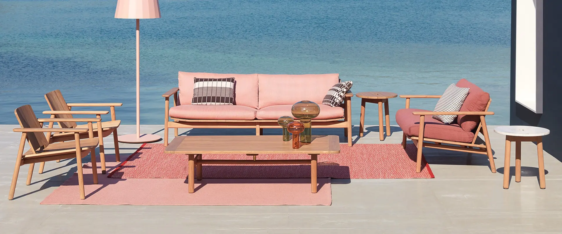 Kettal Riva teak sofa and armchairs with pink cushions on a red rug at the edge of a lake, with green hills behind