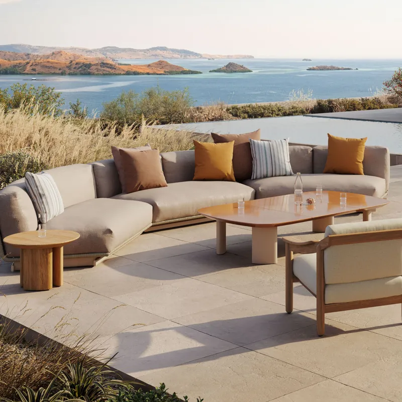 Kettal Tilos curved sofa with striped and ochre cushions beside a teak coffee table on a stone terrace, the sea in the distance