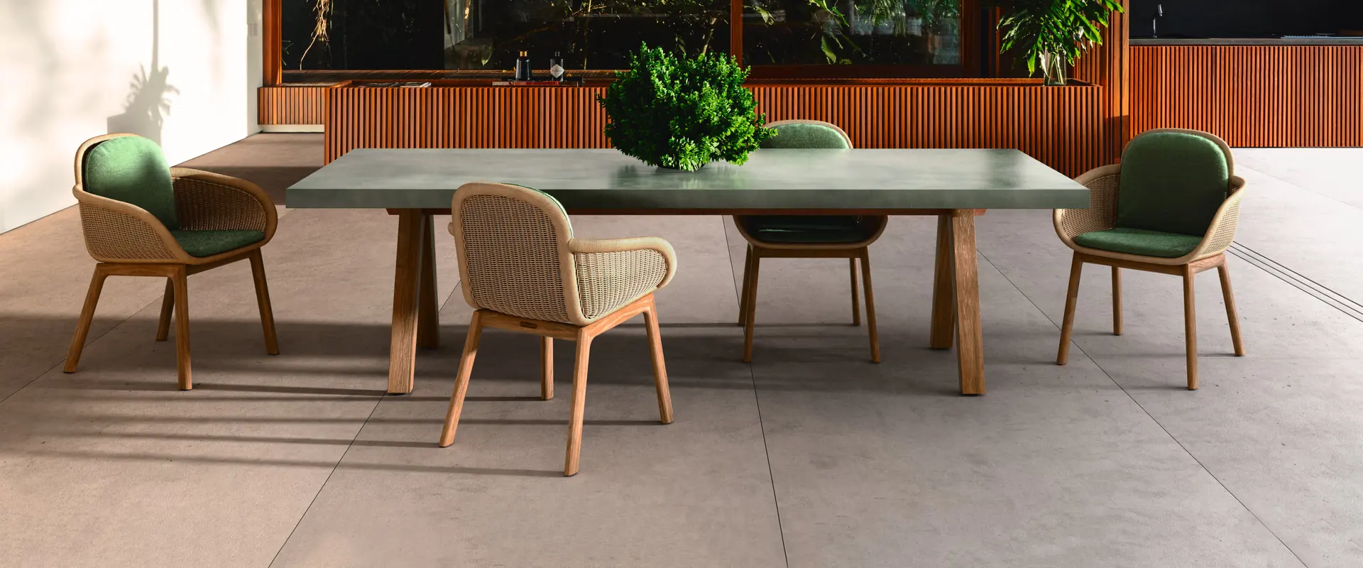 Kettal Vimini wicker dining chairs with green cushions around a stone-topped table on a terrace under a wooden ceiling, forest behind