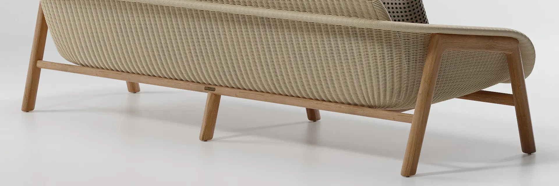 Kettal Vimini three-seater sofa with a woven back and teak legs, seen from behind against a pale studio backdrop
